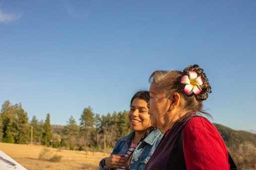 Two women are outdoors, with one smiling broadly while the other looks on with a peaceful expression. The background features a clear blue sky and a landscape with trees and rolling hills. One woman is wearing a denim jacket and the other has a floral hair accessory.