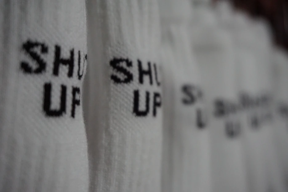 A close-up of a cream-colored t-shirt featuring bold, sarcastic office humor typography in muted charcoal against a washed black background.