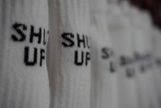 White fabric with black text that repeats 'SHUT UP' in large, bold letters. The image is focused on a close-up of the material, with a blurred background.
