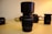 A camera lens with a black hood is placed prominently in the center of the image. The lens, marked with specifications DT 4-5.6/55-200, is surrounded by other photographic equipment, including a partially visible camera and additional lenses in the background. The setup is on a light-colored surface, with warm lighting casting a subtle shadow.