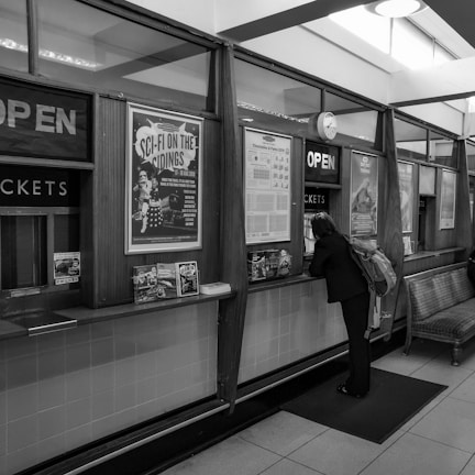 A person stands at a ticket counter in an indoor setting. The walls are adorned with posters and signs, including one promoting a sci-fi event. The counters have 'OPEN' signs and there are some brochures and papers displayed. The lighting is natural from overhead windows and the interior has a vintage feel.