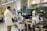 A scientist in a white lab coat and safety goggles is working in a laboratory. The scientist is interacting with a computer and a large, complex piece of lab equipment that is likely used for automated processes. There are several objects present including a cardboard box labeled 'Glass Disposal', various laboratory instruments, and a computer monitor displaying data. The lab is well-lit and organized with cabinets and shelves containing various lab supplies.
