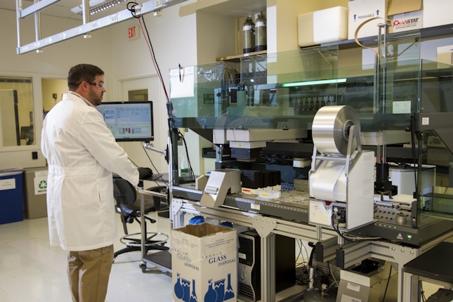 A scientist in a white lab coat and safety goggles is working in a laboratory. The scientist is interacting with a computer and a large, complex piece of lab equipment that is likely used for automated processes. There are several objects present including a cardboard box labeled 'Glass Disposal', various laboratory instruments, and a computer monitor displaying data. The lab is well-lit and organized with cabinets and shelves containing various lab supplies.