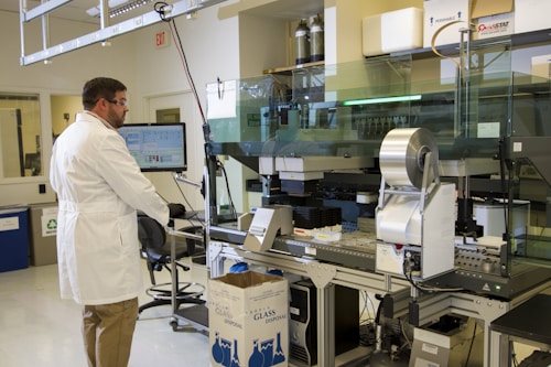 A scientist in a white lab coat and safety goggles is working in a laboratory. The scientist is interacting with a computer and a large, complex piece of lab equipment that is likely used for automated processes. There are several objects present including a cardboard box labeled 'Glass Disposal', various laboratory instruments, and a computer monitor displaying data. The lab is well-lit and organized with cabinets and shelves containing various lab supplies.