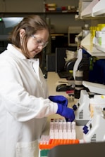A person wearing a white lab coat and blue gloves is handling test tubes in a laboratory setting. The background includes shelves with laboratory equipment and supplies.