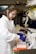 A person wearing a white lab coat and blue gloves is handling test tubes in a laboratory setting. The background includes shelves with laboratory equipment and supplies.
