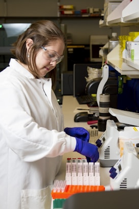 A person wearing a white lab coat and blue gloves is handling test tubes in a laboratory setting. The background includes shelves with laboratory equipment and supplies.