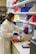 A person wearing a white lab coat and blue gloves is working in a laboratory setting surrounded by various lab equipment. Brightly colored storage boxes in red, yellow, blue, and purple are stacked on shelves. The individual is handling test tubes and operating a piece of laboratory machinery.