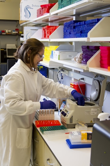 A person wearing a white lab coat and blue gloves is working in a laboratory setting surrounded by various lab equipment. Brightly colored storage boxes in red, yellow, blue, and purple are stacked on shelves. The individual is handling test tubes and operating a piece of laboratory machinery.