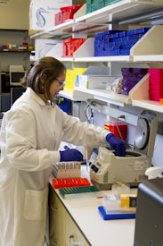A person wearing a white lab coat and blue gloves is working in a laboratory setting surrounded by various lab equipment. Brightly colored storage boxes in red, yellow, blue, and purple are stacked on shelves. The individual is handling test tubes and operating a piece of laboratory machinery.