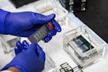 Blue-gloved hands are holding a piece of laboratory equipment, possibly a glass slide with various coatings or samples. The background includes trays filled with liquid and other glass slides.