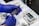 Blue-gloved hands are holding a piece of laboratory equipment, possibly a glass slide with various coatings or samples. The background includes trays filled with liquid and other glass slides.