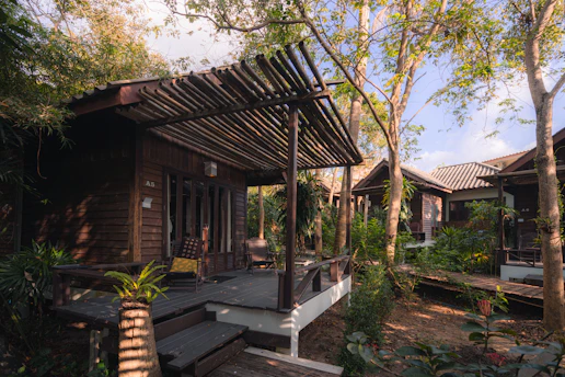 A series of wooden cabins surrounded by lush greenery and trees. The cabins feature wooden decks and pergolas, blending in with the natural environment. Sunlight filters through the trees, creating a serene and tranquil atmosphere. The area appears secluded with a peaceful ambiance, suggesting a retreat or getaway location.