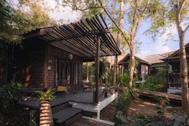 A series of wooden cabins surrounded by lush greenery and trees. The cabins feature wooden decks and pergolas, blending in with the natural environment. Sunlight filters through the trees, creating a serene and tranquil atmosphere. The area appears secluded with a peaceful ambiance, suggesting a retreat or getaway location.