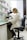 A person wearing a lab coat is seated on a black chair, looking through a microscope on a laboratory bench. The workspace is organized with shelves containing various laboratory supplies, including boxes, bottles, and other equipment. The room is well-lit, and a window with blinds partially open is seen in the background.