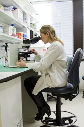 A person wearing a lab coat is seated on a black chair, looking through a microscope on a laboratory bench. The workspace is organized with shelves containing various laboratory supplies, including boxes, bottles, and other equipment. The room is well-lit, and a window with blinds partially open is seen in the background.