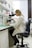 A person wearing a lab coat is seated on a black chair, looking through a microscope on a laboratory bench. The workspace is organized with shelves containing various laboratory supplies, including boxes, bottles, and other equipment. The room is well-lit, and a window with blinds partially open is seen in the background.