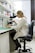 A person wearing a lab coat is seated on a black chair, looking through a microscope on a laboratory bench. The workspace is organized with shelves containing various laboratory supplies, including boxes, bottles, and other equipment. The room is well-lit, and a window with blinds partially open is seen in the background.