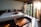 A cozy bedroom with a large bed adorned with decorative pillows and a folded towel in the shape of a swan. Wooden floors and furniture add warmth, while sunlight streams through sheer curtains hanging on a row of tall glass doors.