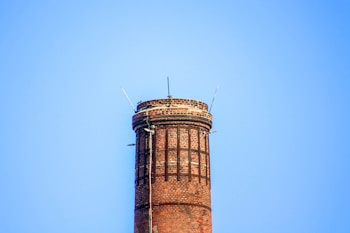 A tall brick chimney stands against a clear blue sky. The chimney is constructed with red bricks and features metal reinforcements. Several metal rods protrude from the top of the chimney.