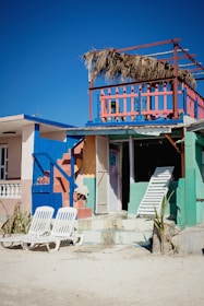 A vibrant beach house with bright colors including blue, pink, and green. The house features a second-story balcony with a straw roof and colorful railing. Two white lounge chairs are placed on the sandy ground in front of the house. A closed door and a leaning white ladder are visible, adding an element of beachside rustic charm.