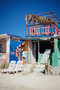 A vibrant beach house with bright colors including blue, pink, and green. The house features a second-story balcony with a straw roof and colorful railing. Two white lounge chairs are placed on the sandy ground in front of the house. A closed door and a leaning white ladder are visible, adding an element of beachside rustic charm.