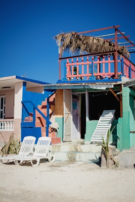 A vibrant beach house with bright colors including blue, pink, and green. The house features a second-story balcony with a straw roof and colorful railing. Two white lounge chairs are placed on the sandy ground in front of the house. A closed door and a leaning white ladder are visible, adding an element of beachside rustic charm.