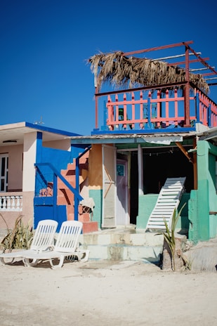 A vibrant beach house with bright colors including blue, pink, and green. The house features a second-story balcony with a straw roof and colorful railing. Two white lounge chairs are placed on the sandy ground in front of the house. A closed door and a leaning white ladder are visible, adding an element of beachside rustic charm.