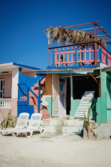 A vibrant beach house with bright colors including blue, pink, and green. The house features a second-story balcony with a straw roof and colorful railing. Two white lounge chairs are placed on the sandy ground in front of the house. A closed door and a leaning white ladder are visible, adding an element of beachside rustic charm.
