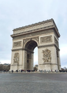 A large triumphal arch with intricate sculptures and carvings stands prominently under a cloudy sky. People gather at the base, emphasizing the monument's grand scale. The structure's detailed artistry highlights historical and cultural significance.