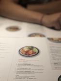 A close-up of a restaurant menu on a table, featuring various ramen dishes. The menu includes detailed descriptions of ingredients such as beef brisket, miso-glazed cod, and kimchee prawns. A blurred image of food can be seen above the text, and a person's arm rests on the table.