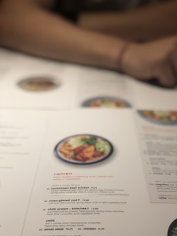 A close-up of a restaurant menu on a table, featuring various ramen dishes. The menu includes detailed descriptions of ingredients such as beef brisket, miso-glazed cod, and kimchee prawns. A blurred image of food can be seen above the text, and a person's arm rests on the table.