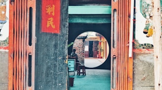 A wooden entrance with traditional Chinese decorations, including vertical red banners with Chinese characters. Beyond the entrance, a round doorway frames a view of an outdoor corridor with brick walls and potted plants. A person is sitting at a small table under a shaded area.