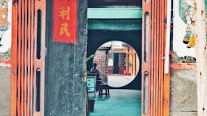 A wooden entrance with traditional Chinese decorations, including vertical red banners with Chinese characters. Beyond the entrance, a round doorway frames a view of an outdoor corridor with brick walls and potted plants. A person is sitting at a small table under a shaded area.