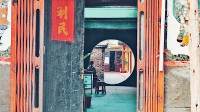 A wooden entrance with traditional Chinese decorations, including vertical red banners with Chinese characters. Beyond the entrance, a round doorway frames a view of an outdoor corridor with brick walls and potted plants. A person is sitting at a small table under a shaded area.