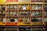 Shelves display rows of glass jars filled with a variety of spices. Each jar has a brass lid and contains different colors and textures, from orange and yellow powders to whole spices and herbs. Above the jars, brightly colored packets are neatly arranged.