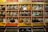 Shelves display rows of glass jars filled with a variety of spices. Each jar has a brass lid and contains different colors and textures, from orange and yellow powders to whole spices and herbs. Above the jars, brightly colored packets are neatly arranged.