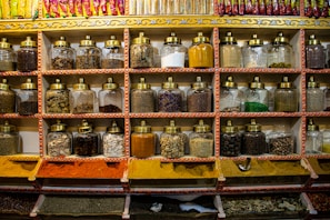 Shelves display rows of glass jars filled with a variety of spices. Each jar has a brass lid and contains different colors and textures, from orange and yellow powders to whole spices and herbs. Above the jars, brightly colored packets are neatly arranged.