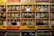 Shelves display rows of glass jars filled with a variety of spices. Each jar has a brass lid and contains different colors and textures, from orange and yellow powders to whole spices and herbs. Above the jars, brightly colored packets are neatly arranged.