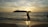 A person standing in a yoga tree pose on a beach during sunset, with the sun positioned behind them creating a silhouette. The ocean waves gently come to shore, and an island is visible in the distance. A parasailer is seen in the sky near the horizon, and there are a few other people in the water.