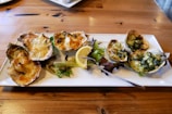 A white rectangular plate on a wooden table holds a neatly arranged serving of baked or grilled oysters topped with melted cheese and herbs. A lemon wedge and some leafy greens garnish the plate. A fork rests on the plate as well.