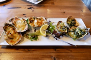 A white rectangular plate on a wooden table holds a neatly arranged serving of baked or grilled oysters topped with melted cheese and herbs. A lemon wedge and some leafy greens garnish the plate. A fork rests on the plate as well.