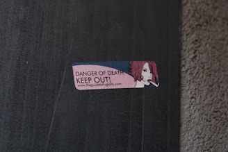 A small rectangular sticker is attached to a concrete wall. The sticker features an illustration of a person with red hair holding a cigarette. The text on the sticker reads 'DANGER OF DEATH KEEP OUT!' along with a URL link.