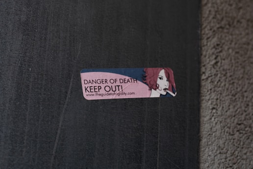 A small rectangular sticker is attached to a concrete wall. The sticker features an illustration of a person with red hair holding a cigarette. The text on the sticker reads 'DANGER OF DEATH KEEP OUT!' along with a URL link.