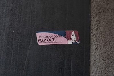 A small rectangular sticker is attached to a concrete wall. The sticker features an illustration of a person with red hair holding a cigarette. The text on the sticker reads 'DANGER OF DEATH KEEP OUT!' along with a URL link.