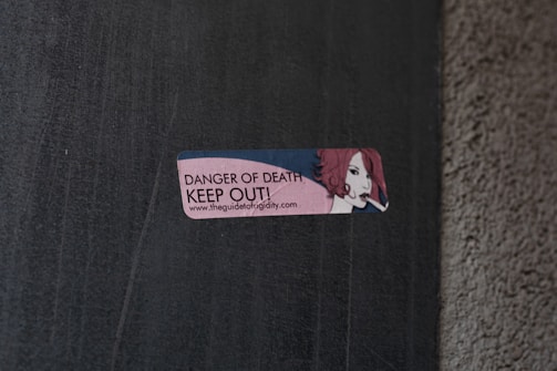 A small rectangular sticker is attached to a concrete wall. The sticker features an illustration of a person with red hair holding a cigarette. The text on the sticker reads 'DANGER OF DEATH KEEP OUT!' along with a URL link.