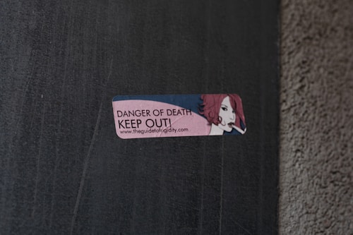 A small rectangular sticker is attached to a concrete wall. The sticker features an illustration of a person with red hair holding a cigarette. The text on the sticker reads 'DANGER OF DEATH KEEP OUT!' along with a URL link.