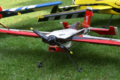 A detailed model airplane with a red, white, and black color scheme is positioned on a grassy surface. The plane features a sleek design with a prominent cockpit, propeller, and landing gear. Another model airplane with a yellow tail and black accents is partially visible in the background.
