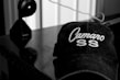 A black and white image featuring a cap with the words 'Camaro SS' embroidered on it, placed on a reflective surface. In the background, a device resembling a camera or webcam is visible with a wire extending across the surface.