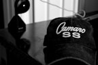 A black and white image featuring a cap with the words 'Camaro SS' embroidered on it, placed on a reflective surface. In the background, a device resembling a camera or webcam is visible with a wire extending across the surface.